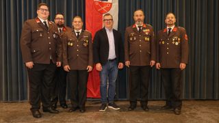 Five men in uniforms stand next to each other in front of a flag. They all look happy.