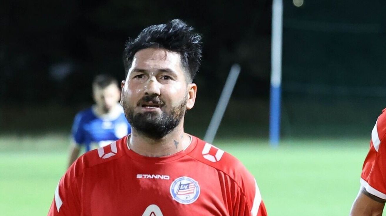 A man in a red jersey stands on a soccer field, looking towards the camera. Behind him, a blurred figure in blue is visible.
