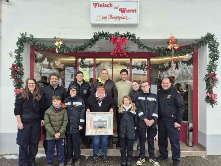 A group of people stand outside a shop named Fleisch und Wurst am Hauptplatz. They hold a framed certificate, dressed in uniforms, and some wear glasses. The shop is decorated with Christmas wreaths.