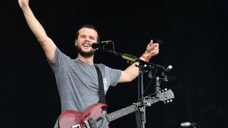 A man in a gray t-shirt sings into a microphone while playing a red guitar on stage.