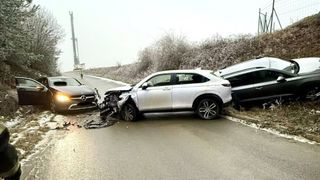 A silver SUV and a black car are involved in a collision on a road, with debris scattered. Nearby, a person stands, and a tall crane is visible in the background.