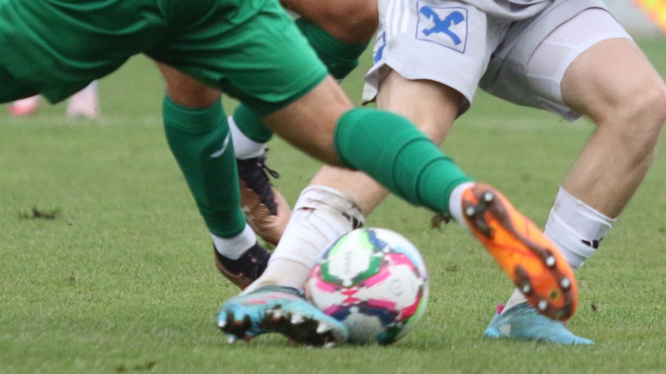 Two soccer players are engaged in a tackle on a green field. One player in green is blocking the other.