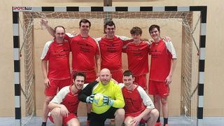A group of men in red jerseys and shorts pose for a photo in front of a net. A man in a yellow vest holds a soccer ball.