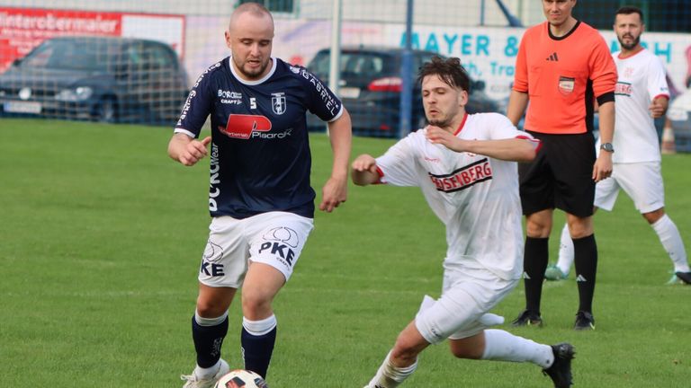 Two soccer players are engaged in a match on a green field. One wears a blue jersey with a sponsor logo, and the other wears a white jersey with red text. Behind them, a blurry background shows parked cars and a fence.