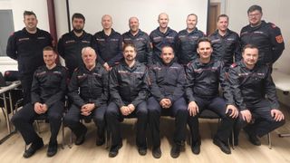 A group of firefighters in uniform pose for a photo, with two rows, one standing and one sitting on chairs, all smiling at the camera.