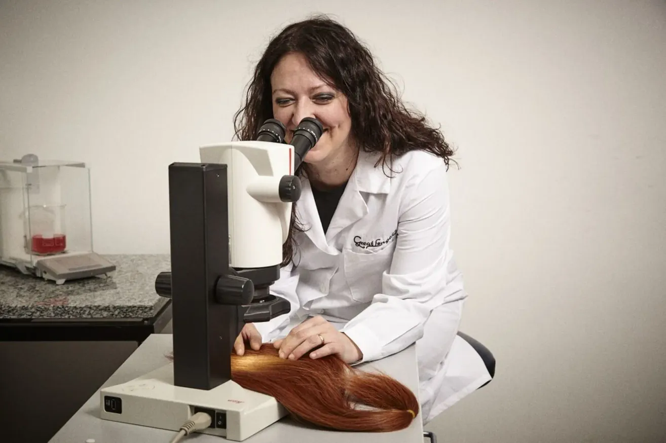 A woman in a lab coat examines long, red hair under a microscope on a table. She is smiling, and the hair is spread out.