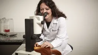 A woman in a lab coat examines long, red hair under a microscope on a table. She is smiling, and the hair is spread out.