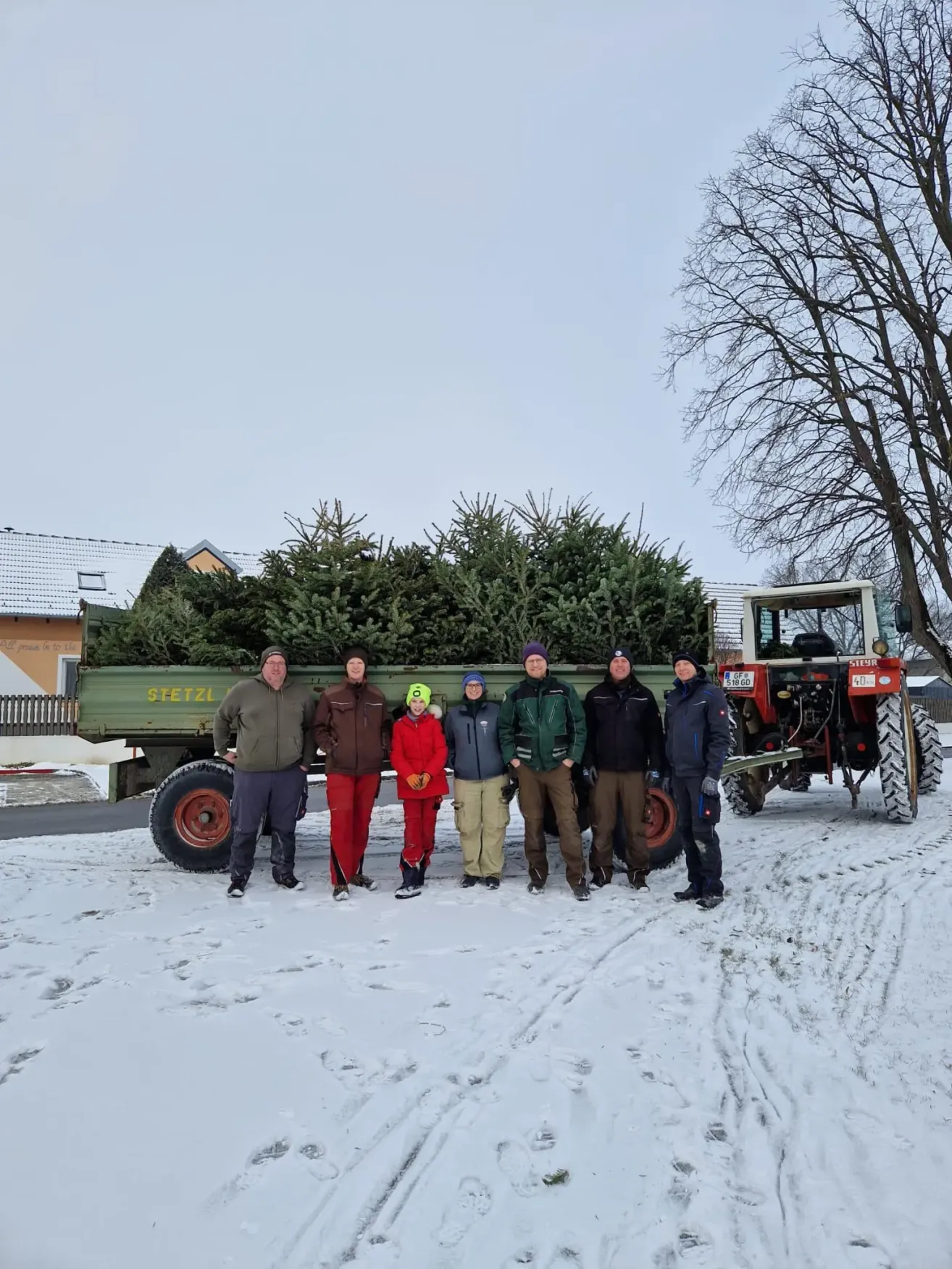 A group of people pose in front of a tractor carrying a load of pine trees. They are bundled in jackets, pants, and hats. Behind them, a house with a slanted roof and a bare tree is visible.