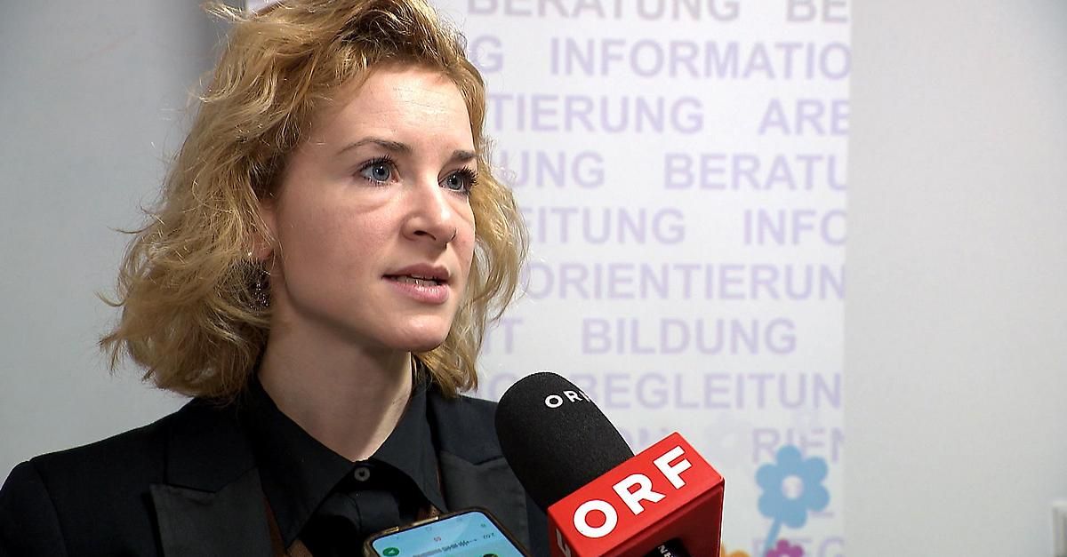A woman with blonde hair is being interviewed by a microphone labeled ORF. She is wearing a black shirt. Behind her, there is a sign with purple text reading 'Bildung,' 'Orientierung,' and 'Begleitung.' There is also a phone in front of the microphone.