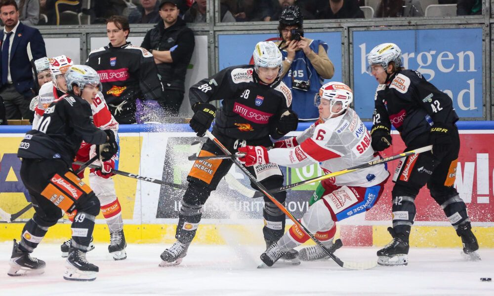 A hockey game with players in action on the ice rink. One player in a black jersey with the number 38 is being checked by two opponents. Spectators are watching from behind the barriers.