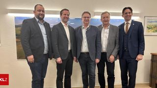 Five men stand in a line, dressed formally in business suits. They all have smiles on their faces. The setting is a room with white walls and a large window in the background.