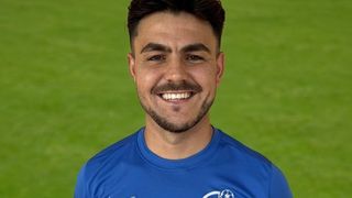 A man in a blue soccer jersey with a Nike logo smiles for a photo on a green field.