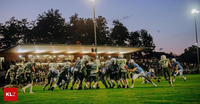 Footballspieler mit Helmen und Trikots spielen auf dem Feld. Im Hintergrund sind Bäume und Straßenlaternen zu sehen.