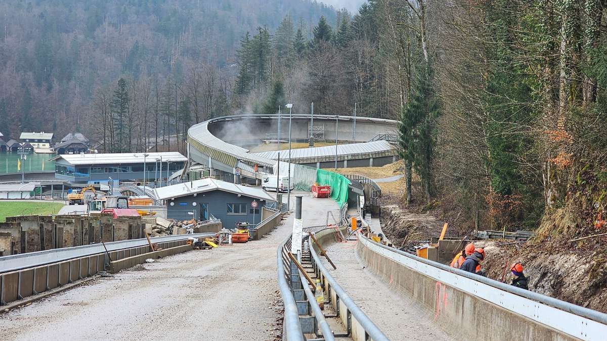 Eine Baustelle mit Arbeitern, mit einer kurvigen Straße, einem blauen Gebäude und einer Metallbarriere. Bäume und Berge im Hintergrund.