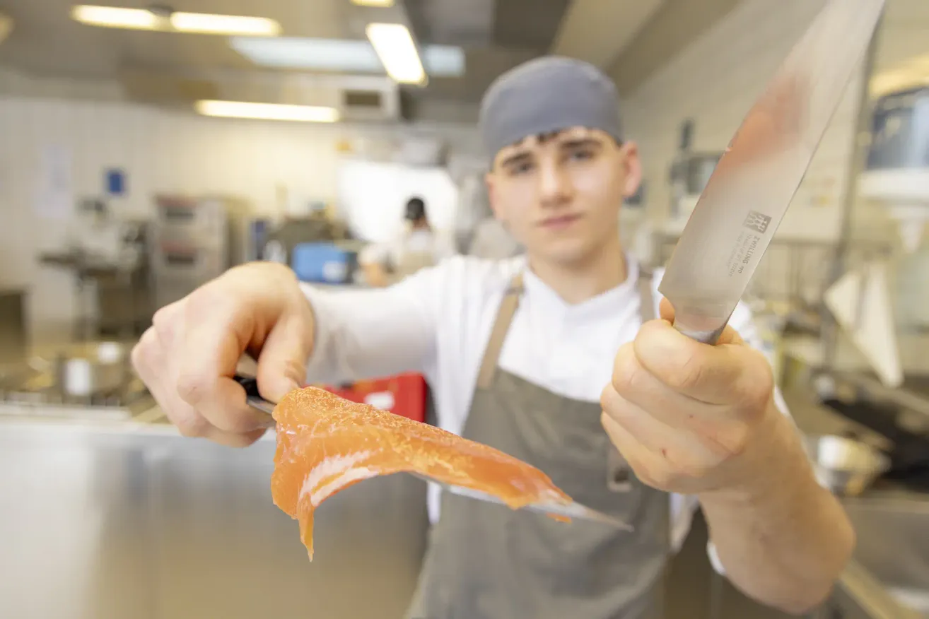 A chef in a kitchen holds a knife, preparing to slice a piece of salmon. He wears a white shirt and a gray apron.