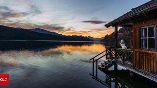 A serene sunset over a calm lake with mountains in the background. A woman stands on a wooden dock, gazing at the sky.