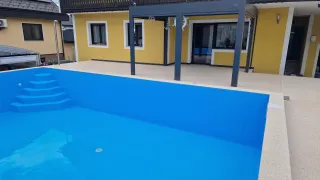 A yellow house with a balcony overlooks a blue swimming pool with a gray pergola. The pool has a drain in the center and steps leading down.