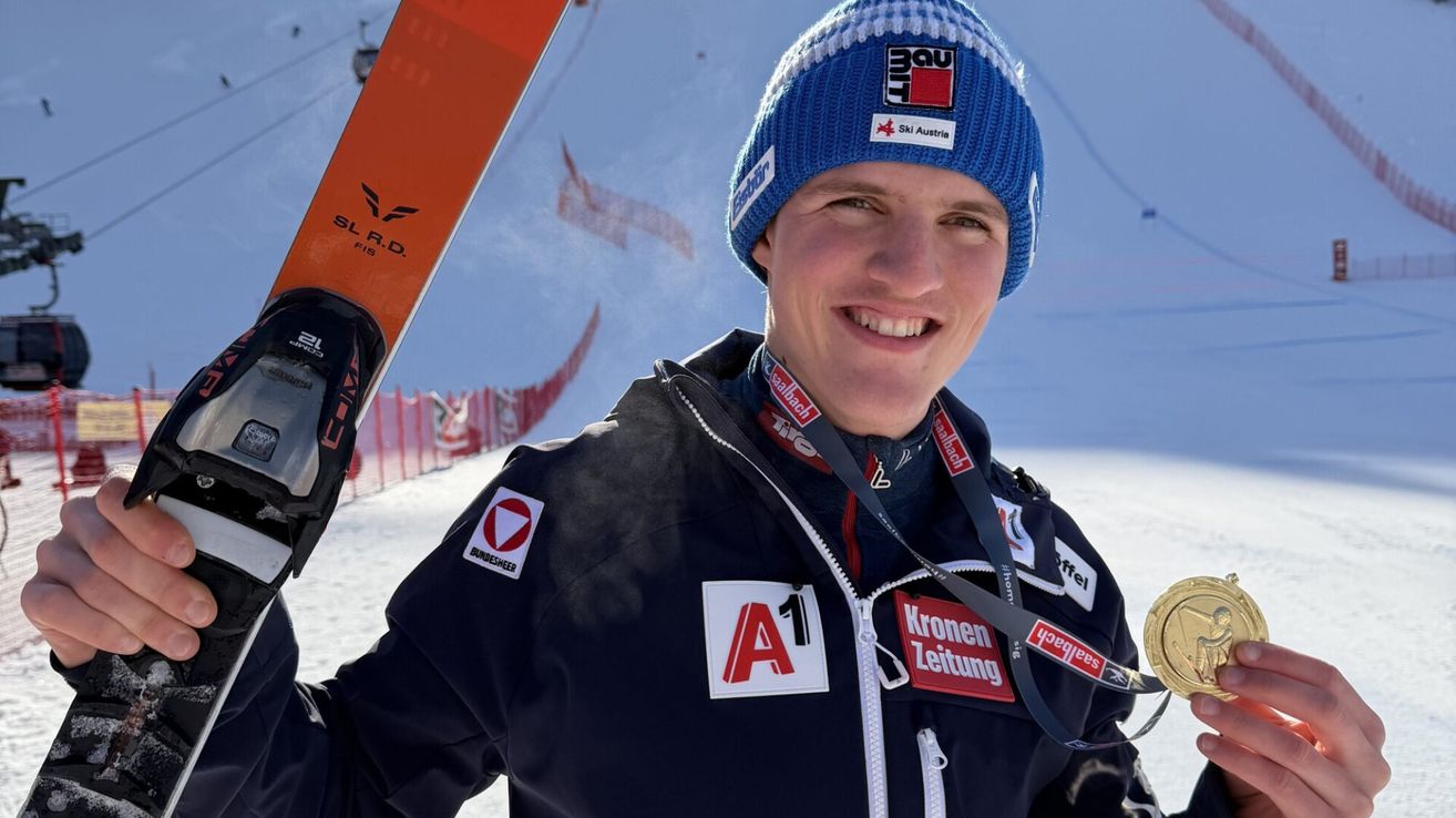 A smiling skier stands in snowy terrain, wearing a blue hat, black jacket, and a silver medal. Behind him is a ski lift and a red barrier.
