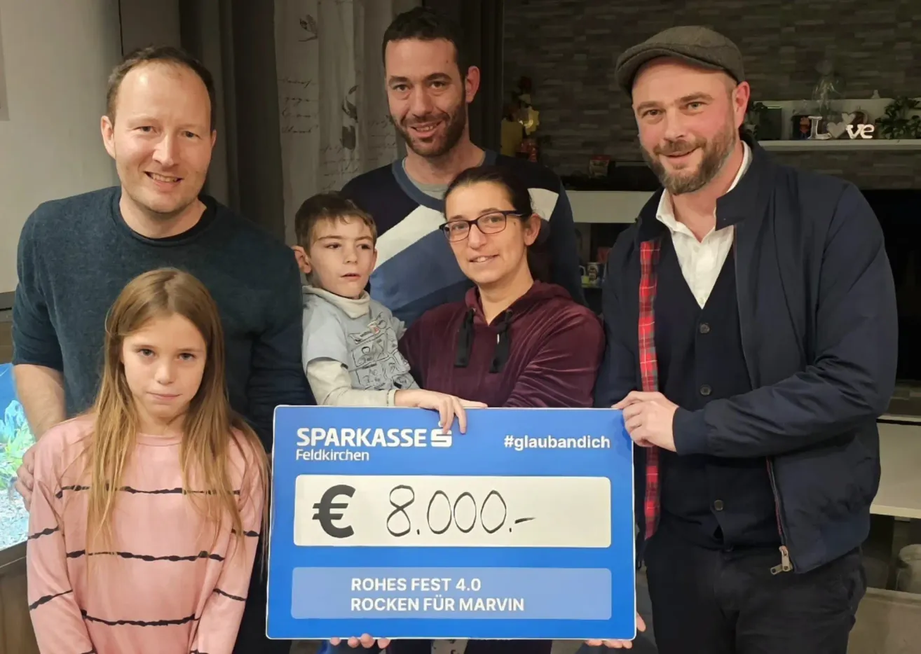 A family of five, including two children, holds a blue sign with the text 'Sparkasse Feldkirchen' and '#glaubandich'. They are smiling and posing for a photo. The boy in the middle is holding a blue sign with the amount '8,000'. Behind them is a blurry background.