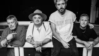 Three men sit together on a bench. The man on the left wears a hat and suspenders. The man in the middle wears a t-shirt with the words 'HOBY DICK'. The man on the right wears a t-shirt and jeans.