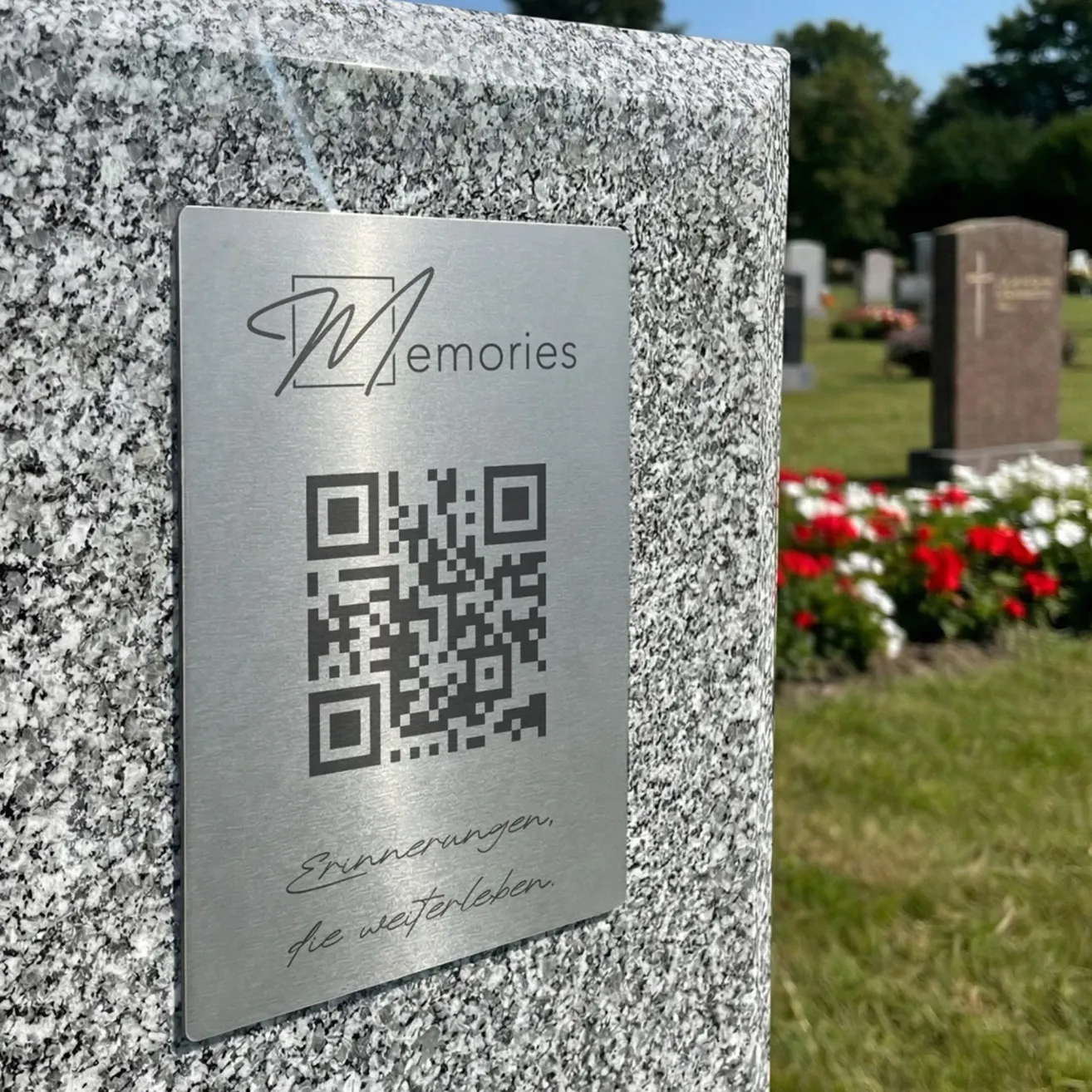 A stainless steel plaque with a QR code and the text 'Memories' is attached to a granite tombstone in a cemetery with flowers.