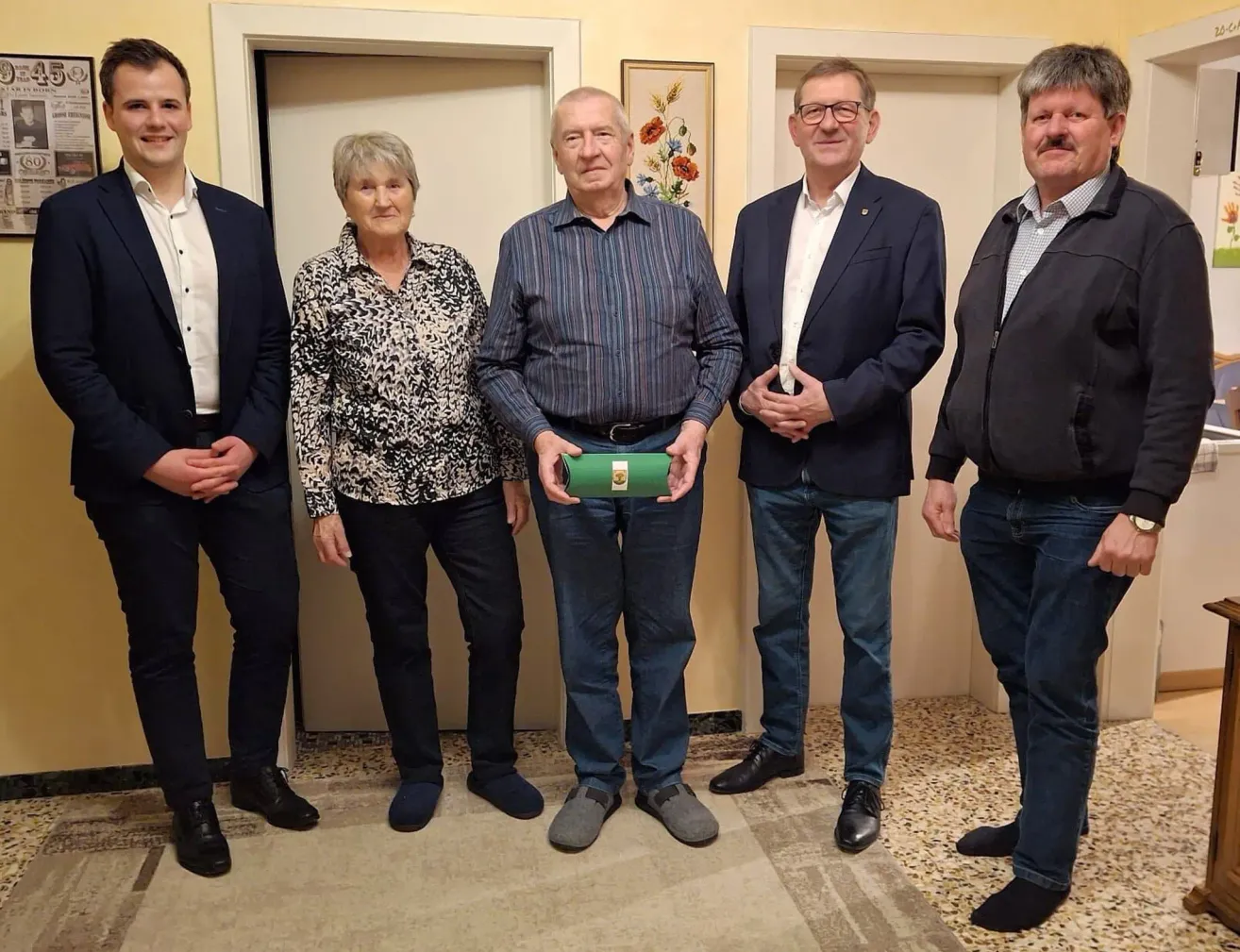 Five individuals stand close together in a room, smiling for a photo. One man holds a green object. Behind them are two white doors.