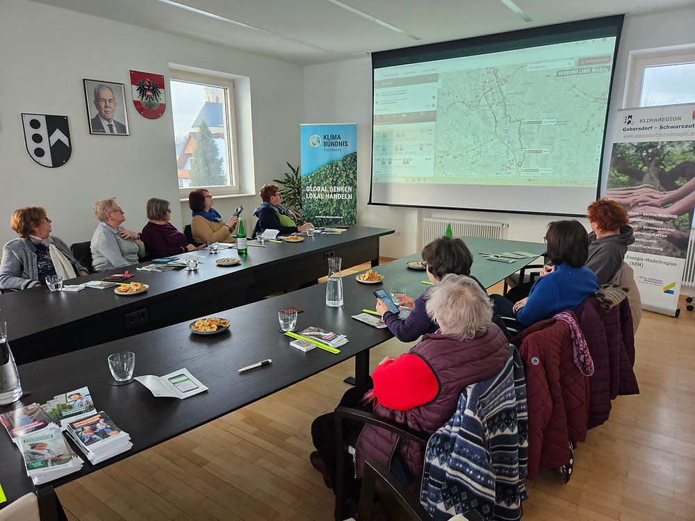 Group of women sit around a table with food and drinks. A presentation on a screen shows a map and text in a foreign language.
