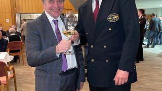 Two men toasting with wine glasses in a banquet hall. One man is wearing a suit and tie, and the other is in a navy jacket with a logo. Behind them, a wooden wall and people seated at tables.