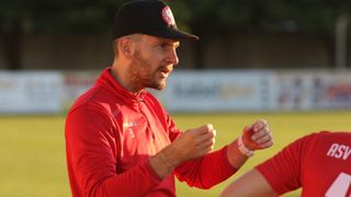 A man in a red jacket and black cap is speaking on a sports field. He wears a watch and a bracelet.