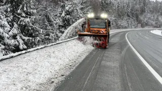 A snowplow clears snow from a road surrounded by snow-covered trees and a distant tower.