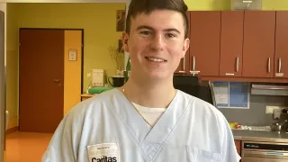 A young man in a white medical coat is smiling in a hospital. He has a name tag that reads Caritas. Behind him is a door, a picture frame, and cabinets.