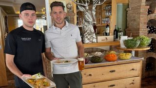 Two men standing in a restaurant holding plates of food. One has a glass of beer. A wooden counter with various dishes and bottles behind them.