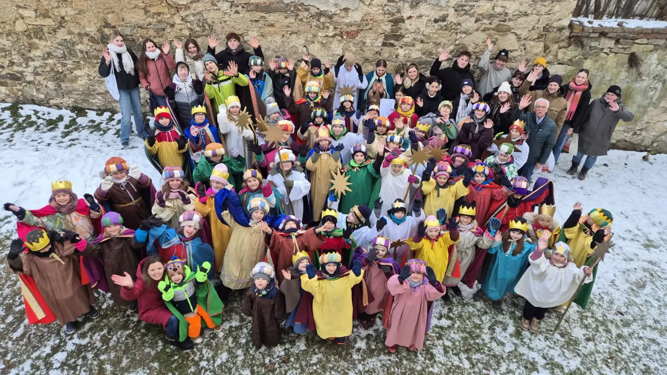 A group of people in festive costumes, some wearing crowns, gather in front of a stone wall, likely for a cultural event. Snow is visible on the ground.