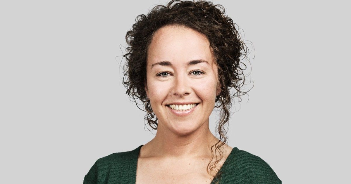 A smiling woman with curly hair wearing a green top poses for a portrait against a gray background.