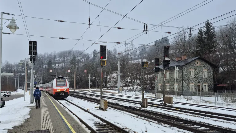 Ein Zug mit roter Signallicht nähert sich dem verschneiten Bahnhof. Schnee bedeckt die Gleise und die Umgebung.