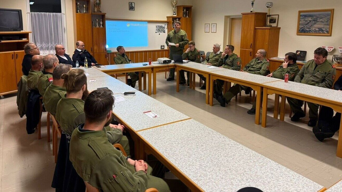 A group of men in military uniforms are seated in a classroom, listening to a man standing in front of a projector screen. A table has a projector and a laptop.