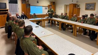 A group of men in military uniforms are seated in a classroom, listening to a man standing in front of a projector screen. A table has a projector and a laptop.