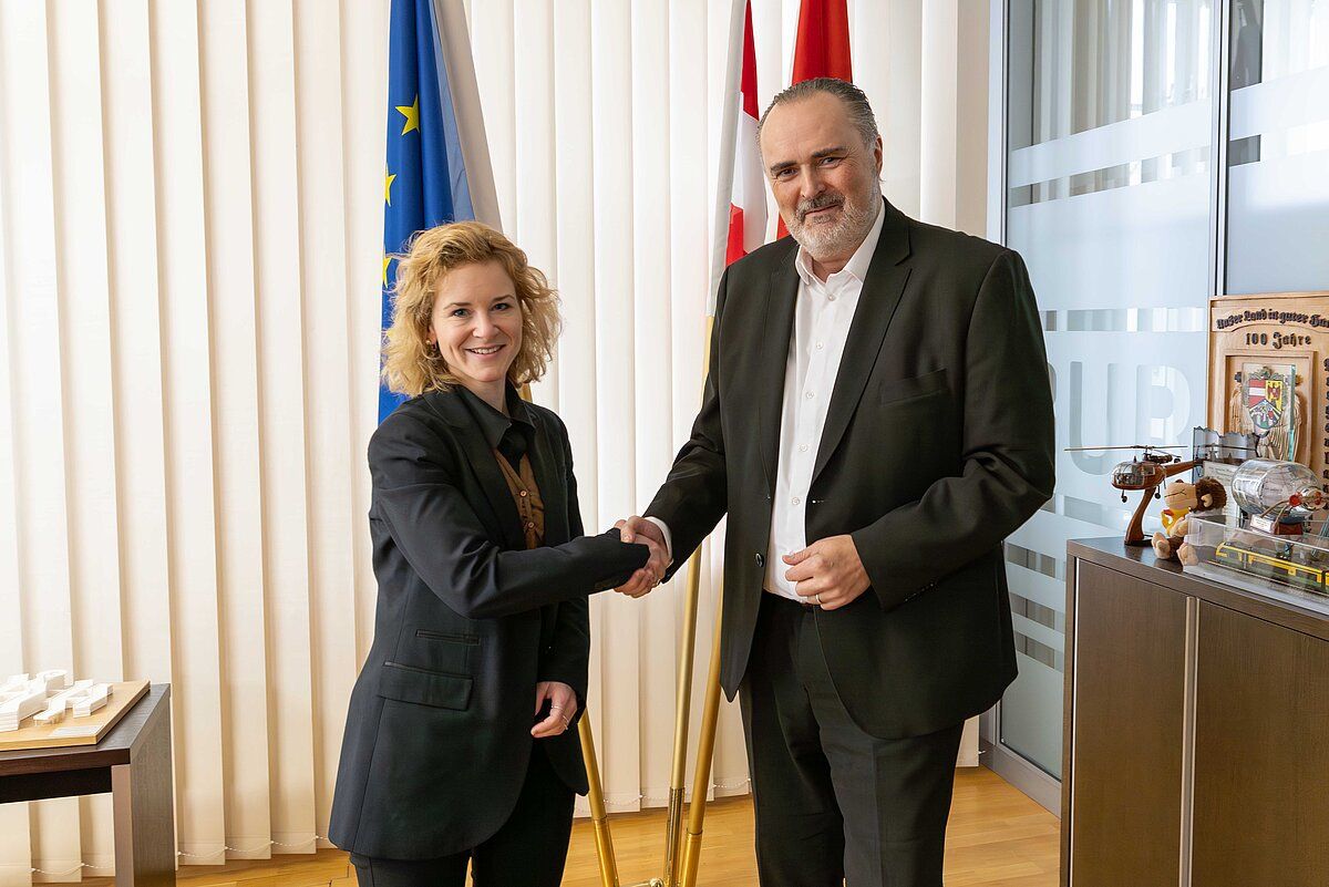 A man and a woman are shaking hands in front of a white wall with flags behind them. The man has a beard and is wearing a dark suit. The woman is smiling and wearing a dark blazer.
