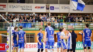 A group of male athletes in blue jerseys stand on a court, likely in a competition. Behind them, a crowd watches from the stands. Banners hang on the wall, one of which reads 'ESIR'.
