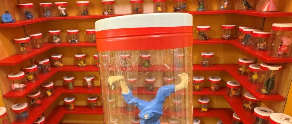 A blue toy figure is in a transparent container with a red cap on a shelf with many other similar containers.