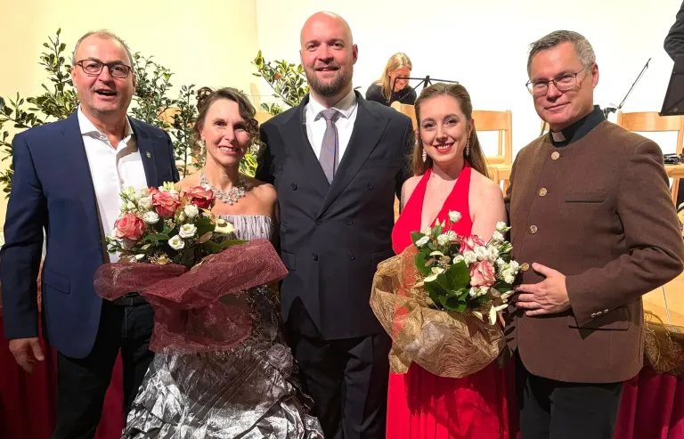 A group of people stand together smiling, possibly at an event. Two women hold bouquets, one in red, the other in silver. A man in formal attire stands between them. In the background, there are chairs and a woman playing an instrument.