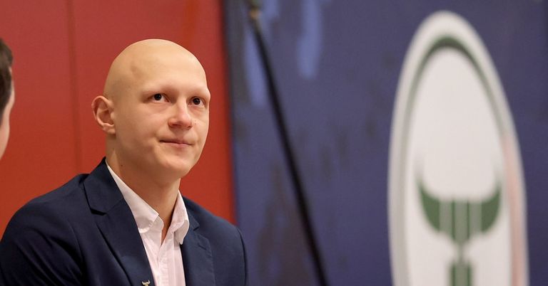A bald man in a navy blue suit and white shirt smiles, standing against a red and blue backdrop with a logo.