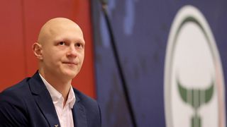 A bald man in a navy blue suit and white shirt smiles, standing against a red and blue backdrop with a logo.