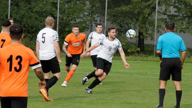 Vier Fußballspieler spielen auf einem Feld. Der Spieler im orangefarbenen Trikot kickt den Ball, während die anderen ihn verfolgen. Bäume und ein Zaun sind im Hintergrund zu sehen.