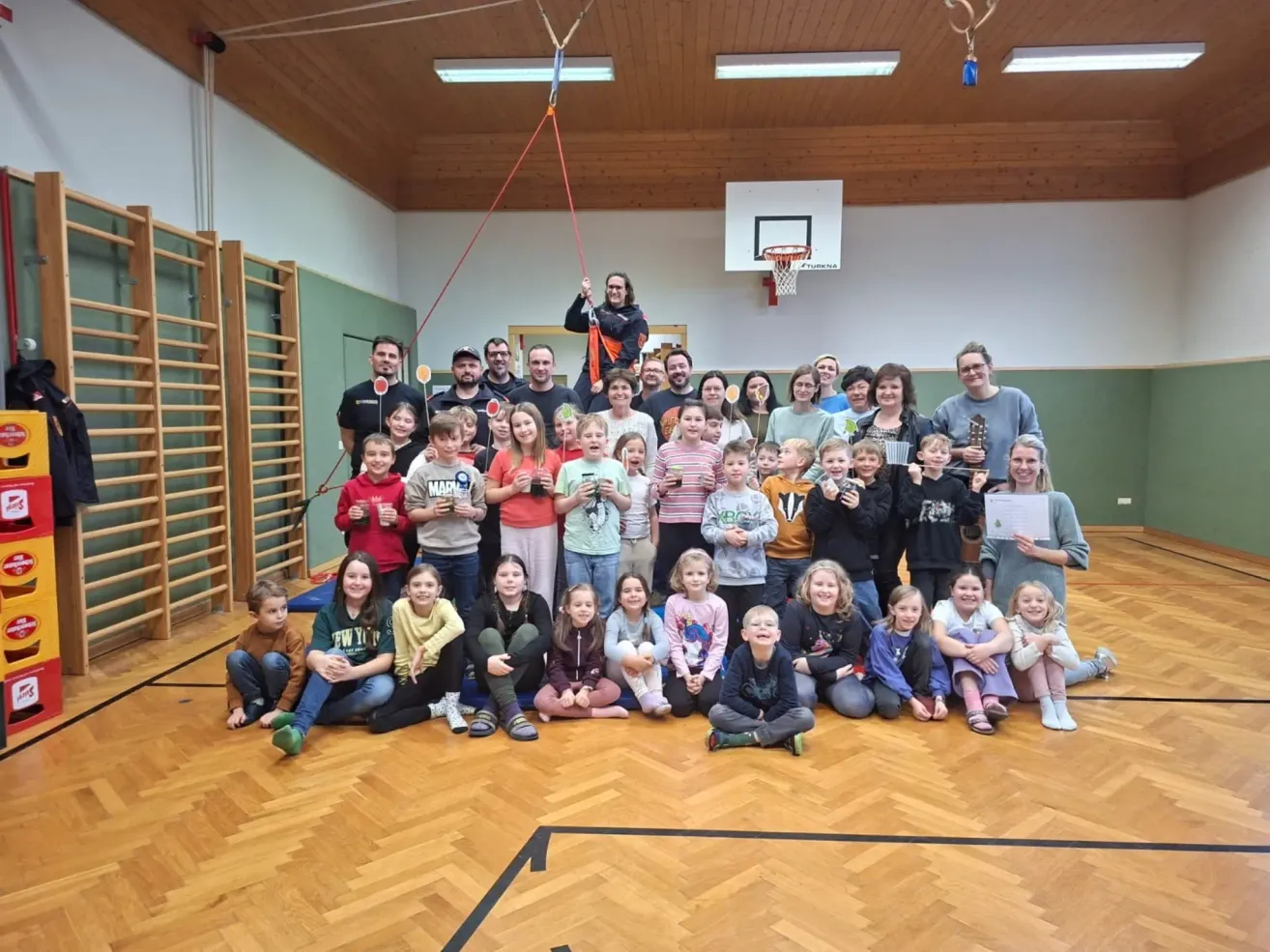A group of children and adults are posing for a picture in a gym with a basketball hoop. A person is holding a rope from the ceiling.