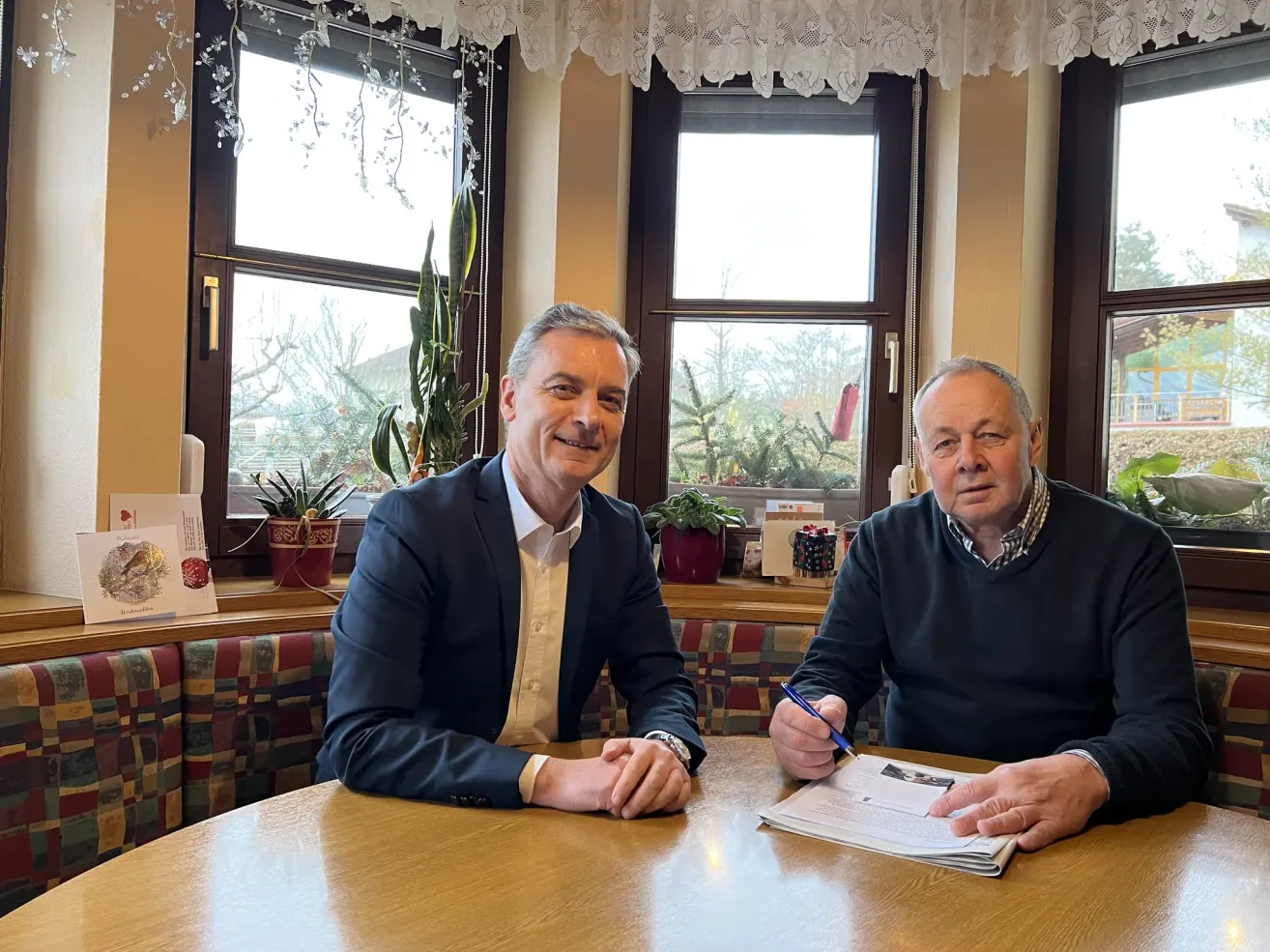 Zwei Männer sitzen an einem Tisch in einem Restaurant. Einer trägt einen Anzug und der andere einen Pullover. Sie lächeln beide und scheinen ein Dokument zu unterschreiben. Auf der Fensterbank stehen Blumentöpfe.