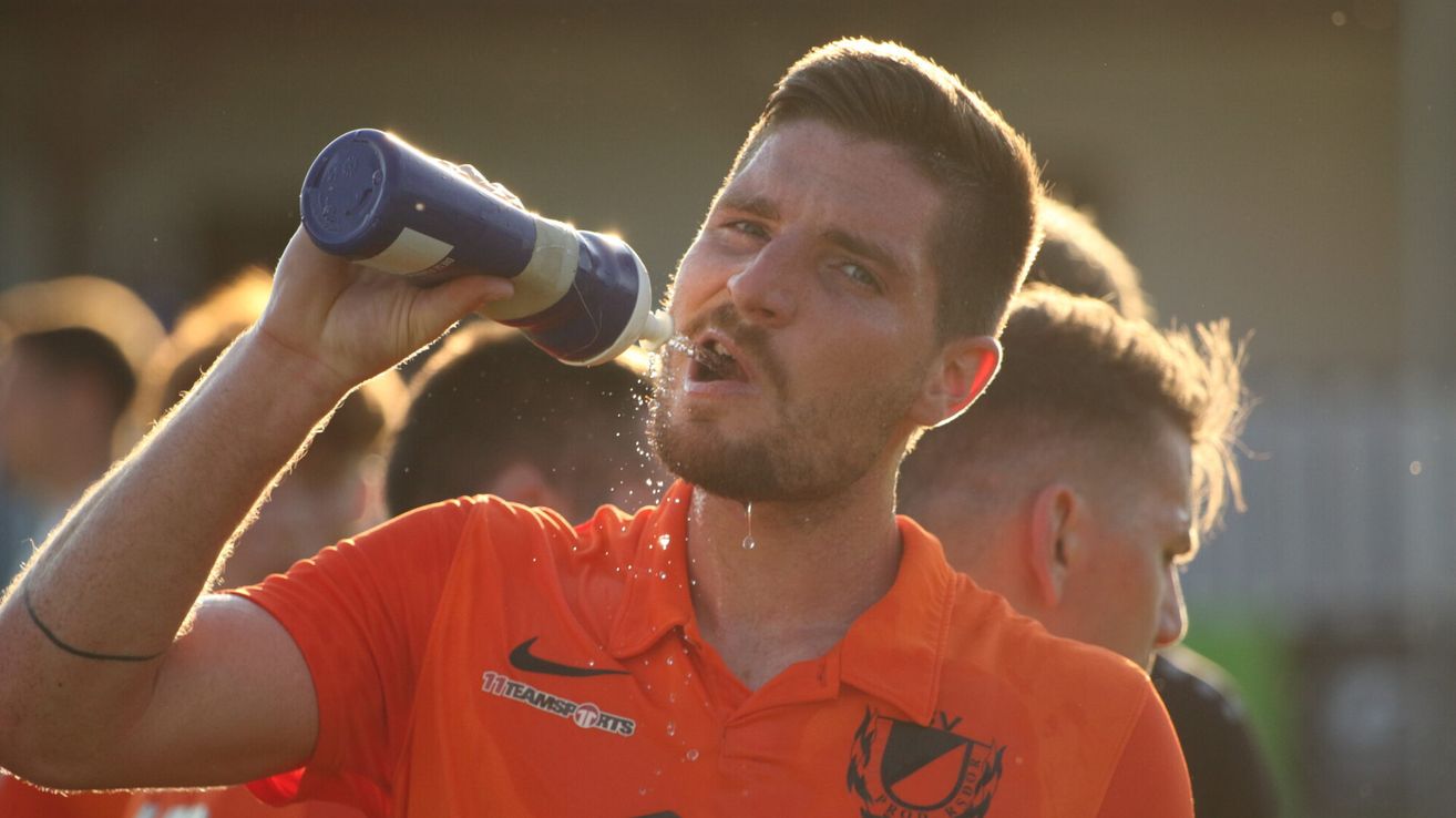 A man drinks from a blue and white bottle, with water dripping from his mouth. He wears an orange shirt with a logo on the left side. Blurry people are behind him.