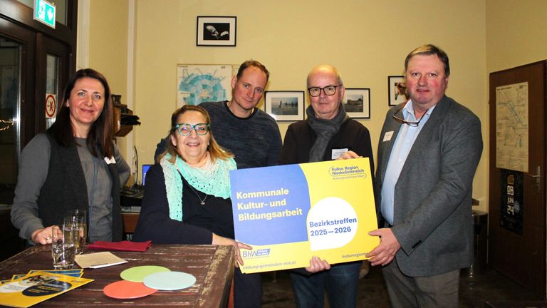 Vier Personen posieren für ein Foto in einem Raum. Eine Frau hält ein gelbes und blaues Schild mit der Aufschrift 'Kommunale Kultur- und Bildungsarbeit Bezirkstreffen 2025-2026'. Der Raum hat gelbe Wände und gerahmte Fotos.
