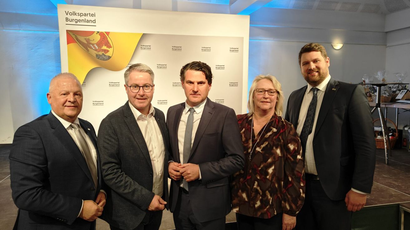 Fünf Menschen in formeller Kleidung posieren für ein Foto vor einer Wand mit Logo und Text.