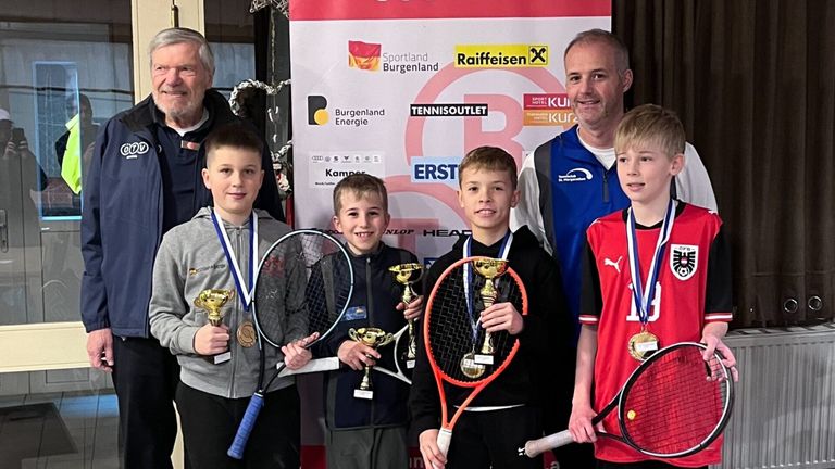 Vier Jungen, die Tennisschläger und Medaillen halten, stehen vor einem Banner mit verschiedenen Sponsorenlogos. Ein älterer Mann mit Bart steht hinter ihnen.